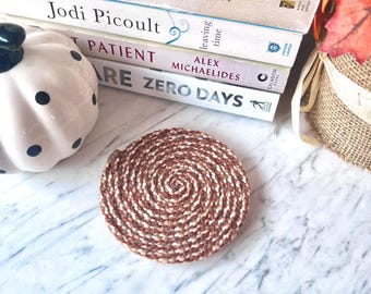 May include: A round coaster featuring a spiral pattern of braided fibers in shades of brown, cream, and tan. The coaster is positioned on a white marble surface near a white ceramic pumpkin with black polka dots and a stack of hardcover books.