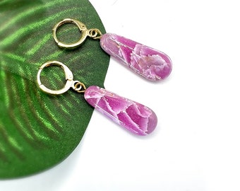 May include: A pair of dangle earrings with gold-colored leverback closures. The earrings feature teardrop-shaped, pink and white marbled stones. The earrings are displayed on a green leaf against a white background.