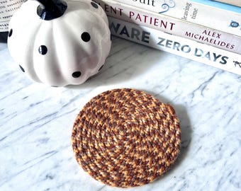 May include: A round coaster crafted from braided fabric in a variegated pattern of brown, tan, and orange tones. The coaster features a tight, circular coil construction with a visible spiral texture throughout the entire surface of the piece.