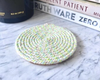 May include: A circular coaster constructed from coiled rope featuring a variegated color palette of soft beige, light mint green, and pale yellow. The rope is tightly wound in a spiral pattern, creating a textured surface with a consistent thickness.