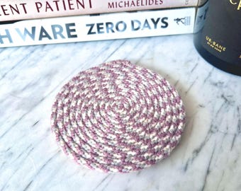 May include: A circular coaster crafted from coiled rope in a variegated pattern of mauve, white, and grey. The rope is tightly wound in a spiral design, creating a textured surface with a consistent thickness throughout the round shape.