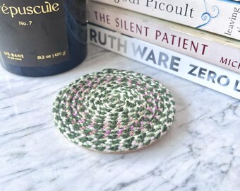 May include: A round, braided coaster featuring a variegated pattern of dark green, light green, and soft pink yarn fibers. The coaster is constructed from a continuous coiled rope design, creating a textured, circular surface for resting items.