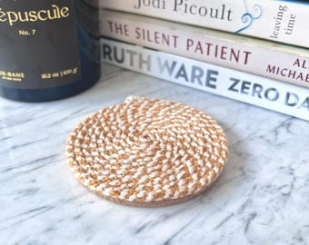 May include: A circular coaster featuring a spiral pattern of intertwined white and tan fibers. The coaster has a flat, round shape with a textured surface created by the coiled material, providing a neutral color palette and a consistent woven design.