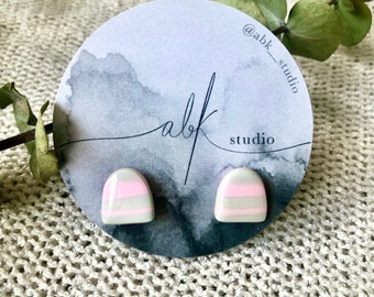 May include: A pair of stud earrings with a rounded top design. The earrings are white with horizontal stripes of pink and gray. They are displayed on a round card with the text "abk studio".