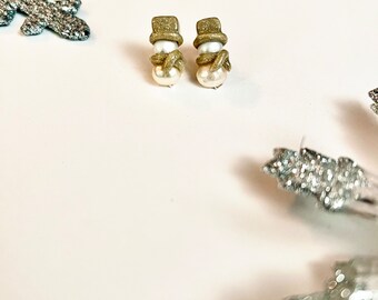 May include: A pair of snowman earrings. Each earring features a white pearl body, a gold glitter top hat, and a gold scarf. The earrings are set against a white background with silver snowflake decorations.