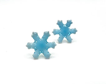 May include: Two light blue snowflake-shaped earrings. Each earring has six points and a slightly textured surface. The earrings are set against a white background, highlighting their delicate design. These earrings are perfect for winter or holiday wear.
