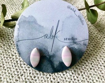 May include: A pair of oval-shaped stud earrings with a pink and white striped design. The earrings are displayed on a round card with the text "abk studio" and the Instagram handle "@abk_studio". The background is a watercolor design.