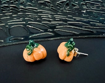 May include: A pair of orange pumpkin-shaped earrings with green stems. The earrings are set against a dark background, creating a contrast that highlights the autumnal theme. The earrings are small and detailed.