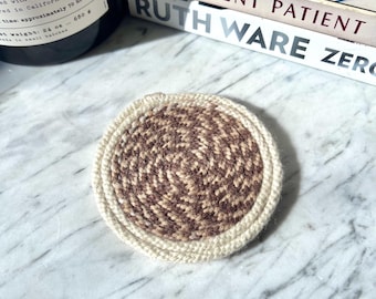 May include: A circular crochet coaster featuring a central spiral pattern in alternating shades of dark brown and light brown yarn, surrounded by a thick, solid cream-colored border. The coaster is crafted with a visible, textured stitch pattern.