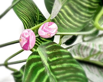 May include: Two oval-shaped stud earrings with pink floral designs. The earrings are set against a backdrop of green leaves and vines. The earrings have a light pink base color with darker pink floral patterns.