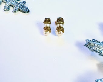 May include: A pair of snowman-shaped earrings. The earrings feature a gold glitter hat and scarf, with a white pearl body. The earrings are set against a white background with silver glitter snowflake accents.