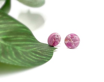 May include: Two round, pink stud earrings with a marbled appearance. The earrings have a silver post and are set against a white background with a green leaf in the corner. The earrings are a jewelry item.