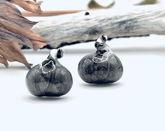 May include: Two small, dark gray ceramic fig-shaped sculptures with a glossy finish. Each has a white and gray swirl detail on top. The figs are set against a white background with a piece of driftwood and dried plant matter.