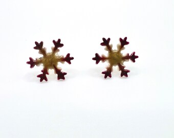 May include: A pair of snowflake-shaped stud earrings. The earrings have a gold-colored center with burgundy-colored edges. The earrings are set against a white background.