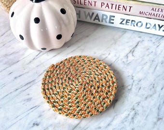 May include: A round coaster crafted from coiled rope in a variegated pattern of orange, green, and cream fibers. The coaster features a tight, circular weave that creates a textured surface, showcasing a blend of autumnal colors throughout the design.