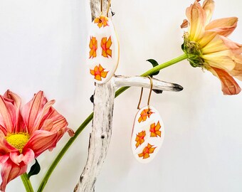 May include: White oval earrings with a floral design in shades of orange and pink, suspended from gold-colored hooks. The earrings are displayed on a piece of driftwood, with a pink and yellow flower in the background. The earrings are a unique accessory.