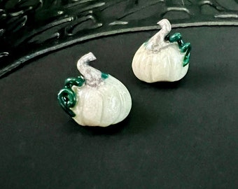 May include: Two small, white pumpkin-shaped ornaments with gray stems and green vines. The pumpkins are set against a dark background, creating a contrast that highlights their shape and color. These decorative items are likely for fall or Halloween.