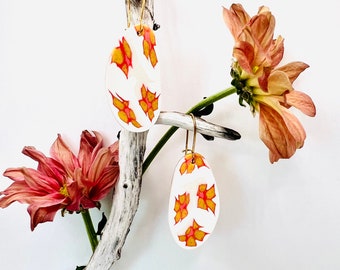 May include: Oval-shaped earrings with a white background and a pattern of orange and red leaf-like shapes. The earrings hang from gold-colored hooks. The earrings are displayed on a white branch with dried flowers.