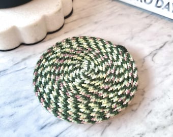 May include: A circular coaster featuring a dense, spiraling knit pattern in variegated shades of forest green, pale yellow, and soft pink. The yarn is tightly coiled to create a flat, round surface with a textured, multi-colored appearance.