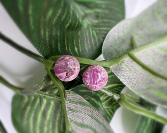 May include: Two round, pink earrings with a marbled texture. The earrings are set against a backdrop of green leaves with white accents, creating a natural and elegant presentation. The earrings are a perfect accessory for any outfit.