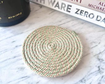 May include: A circular coaster crafted from a continuous coil of braided rope in alternating shades of sage green and soft beige. The rope is tightly wound in a spiral pattern, creating a textured, flat surface with a small loop at the edge.