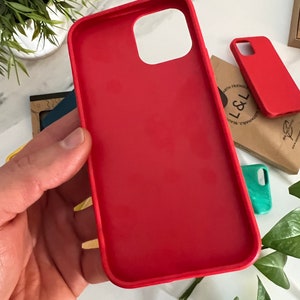 May include: A vibrant red phone case, held in a hand, showcasing a smooth, matte finish. The case has precise cutouts for the camera and buttons. Additional red and teal cases are visible in the background, along with a small plant and a brown paper bag.