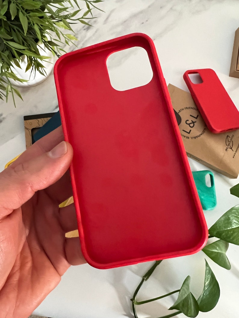 May include: A vibrant red phone case, held in a hand, showcasing a smooth, matte finish. The case has precise cutouts for the camera and buttons. Additional red and teal cases are visible in the background, along with a small plant and a brown paper bag.