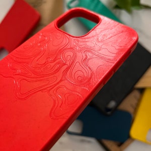 May include: A close-up of a red phone case with a topographic map design. Other phone cases in teal, black, and yellow are visible in the background. The case has a smooth texture and a cutout for the camera.