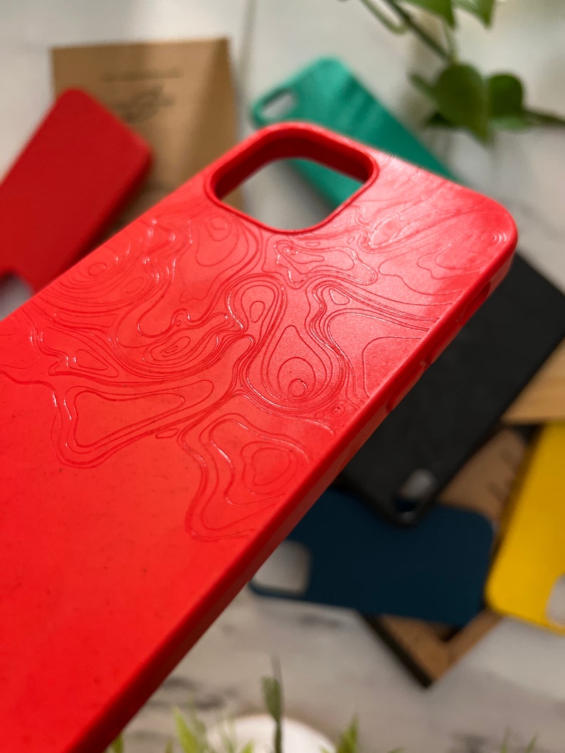 May include: A close-up of a red phone case with a topographic map design. Other phone cases in teal, black, and yellow are visible in the background. The case has a smooth texture and a cutout for the camera.