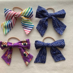 May include: Four handmade hair bows with different patterns. The bows are made of fabric and have a simple design. The bows are in various colors, including purple, blue, and white.