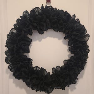 K&ouml;nnte beinhalten: Ein runder, schwarzer Jute-Kranz mit einem R&uuml;schendesign. Der Kranz besteht aus einem gewebten Material und h&auml;ngt an einer wei&szlig;en T&uuml;r. Der Kranz ist ein dekorativer Gegenstand.