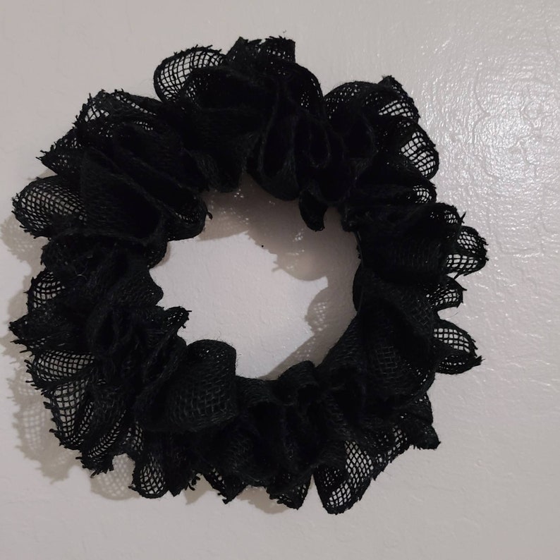 K&ouml;nnte beinhalten: Ein schwarzes Haargummi aus ger&uuml;schtem, netzartigem Stoff. Das Haargummi ist kreisf&ouml;rmig und scheint handgefertigt zu sein. Die dunkle Farbe und Textur deuten auf eine gotische oder kantige &Auml;sthetik hin, die f&uuml;r verschiedene Frisuren geeignet ist.