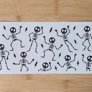 May include: A sheet of black and white skeleton stickers with a lightning bolt design. The skeletons are in various poses, some are dancing, some are holding their arms up, and some are holding their arms down.