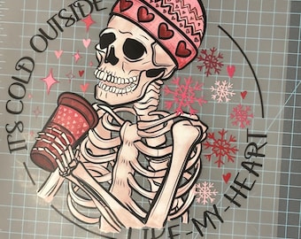 May include: A pink and white skeleton wearing a pink knit hat with hearts and holding a pink and white coffee cup. The skeleton is surrounded by snowflakes and hearts. The text "It's cold outside like my heart" is written around the skeleton.
