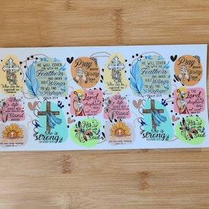 May include: A sheet of colorful stickers with inspirational quotes and bible verses. The stickers feature a variety of designs, including crosses, flowers, and birds. The text on the stickers includes phrases like "She is strong", "Pray more worry less", and "He will cover you with his feathers".