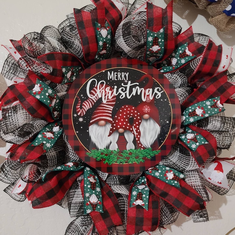 Puede incluir: Una corona navide&ntilde;a festiva con un letrero redondo central con las palabras "Merry Christmas" e ilustraciones de tres gnomos. La corona est&aacute; adornada con cintas y detalles a cuadros rojos y negros.