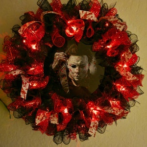 Puede incluir: Una corona de Halloween con malla negra y roja, cinta blanca y luces rojas. Una imagen de Michael Myers de la película Halloween está en el centro de la corona.