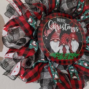 Puede incluir: Una corona navide&ntilde;a festiva con una pieza central circular que presenta tres gnomos y las palabras "Merry Christmas". La corona incorpora cinta de cuadros de b&uacute;falo roja y negra, cinta verde con patrones de gnomos y copos de nieve y malla blanca.