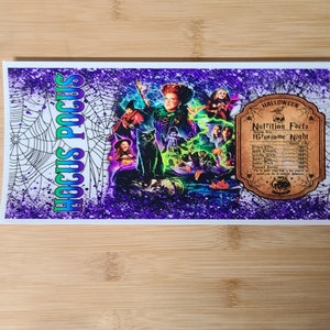 May include: A rectangular sticker with a Halloween theme. The sticker features the text "Hocus Pocus" in green and white, a graphic of characters from the movie, and a "Nutrition Facts" label. The background is purple with a spiderweb design.