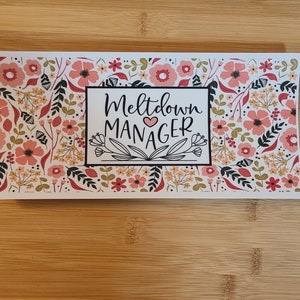 May include: White sticker with a floral pattern in pink and green. The sticker has black text that reads "Meltdown Manager" with a small pink heart.