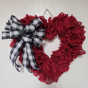 Puede incluir: Una corona de corazón de arpillera roja con un lazo a cuadros blanco y negro. La corona está decorada con volantes y es perfecta para el Día de San Valentín o cualquier ocasión.