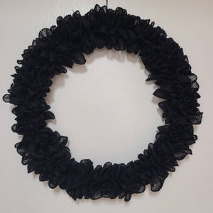 K&ouml;nnte beinhalten: Ein kreisf&ouml;rmiger schwarzer Kranz mit ger&uuml;schter Textur. Der Kranz besteht aus einem dunklen, m&ouml;glicherweise leinenartigen Material und wird vor einem schlichten wei&szlig;en Hintergrund pr&auml;sentiert. Das Design ist schlicht und elegant.
