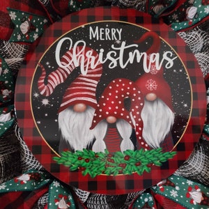 Puede incluir: Una corona navide&ntilde;a con un letrero redondo central que muestra tres ilustraciones de gnomos con sombreros rojos. El letrero dice "Merry Christmas" sobre un fondo negro con un borde a cuadros rojo y negro. Cintas y lazos verdes y rojos completan el dise&ntilde;o.