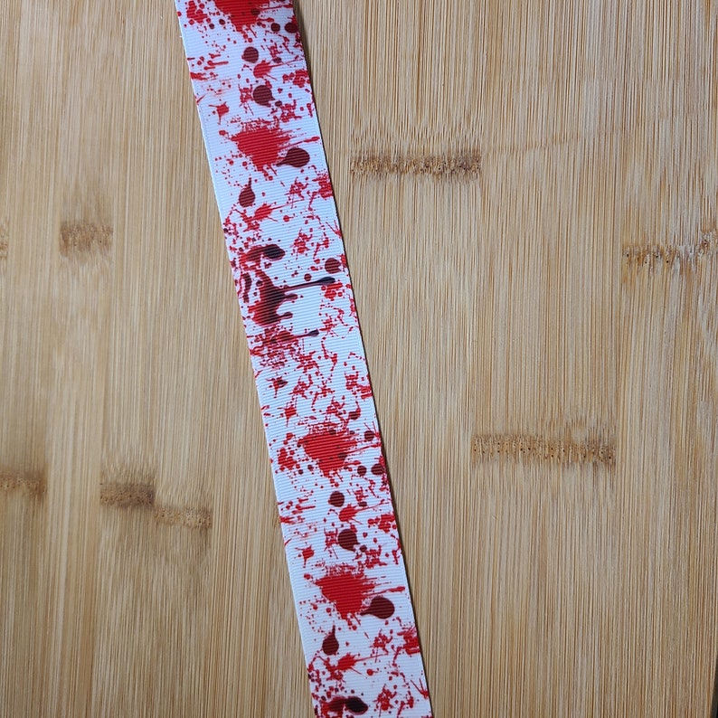 May include: White ribbon with red blood splatter design. The ribbon is on a light brown wooden surface.