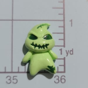 May include: A green and black plastic figurine of a ghost with a stitched mouth and green eyes. The ghost is standing on a white surface with a ruler in the background.