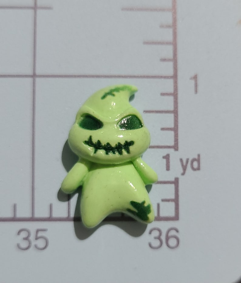 May include: A green and black plastic figurine of a ghost with a stitched mouth and green eyes. The ghost is standing on a white surface with a ruler in the background.