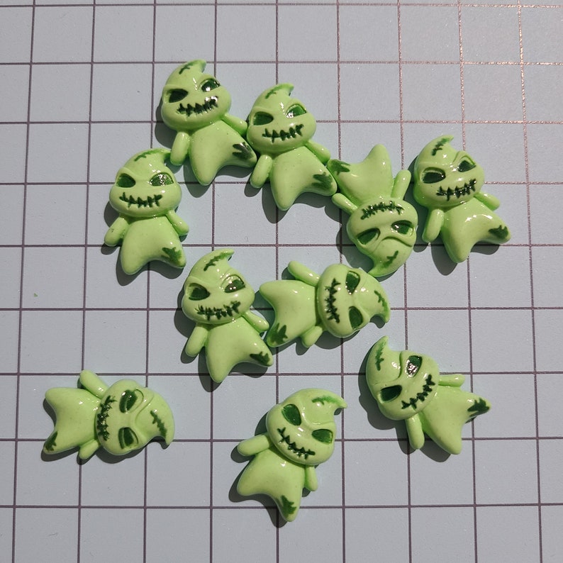 May include: Ten green plastic Halloween character charms with stitched-up mouths and green eyes. The charms are shaped like a ghost with pointy ears and a rounded body.