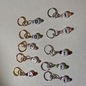 May include: A collection of handmade keychains with letter beads. Each keychain features a small, round bead with the letter "D" or the number "1" on it, along with colorful accent beads and a metal keyring. The keychains are in various colors.