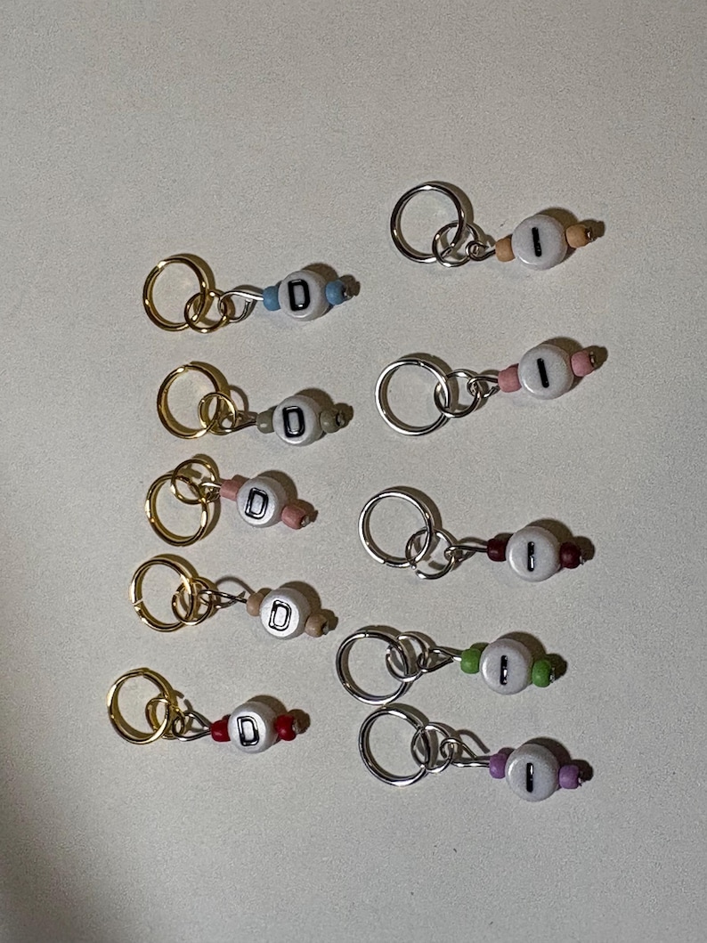 May include: A collection of handmade keychains with letter beads. Each keychain features a small, round bead with the letter "D" or the number "1" on it, along with colorful accent beads and a metal keyring. The keychains are in various colors.