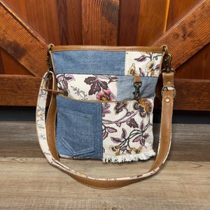 May include: A patchwork crossbody bag with a denim pocket and floral print panels. The bag features a brown leather strap and trim, with a fringed edge on one panel. The bag has a brown leather strap and a denim pocket.